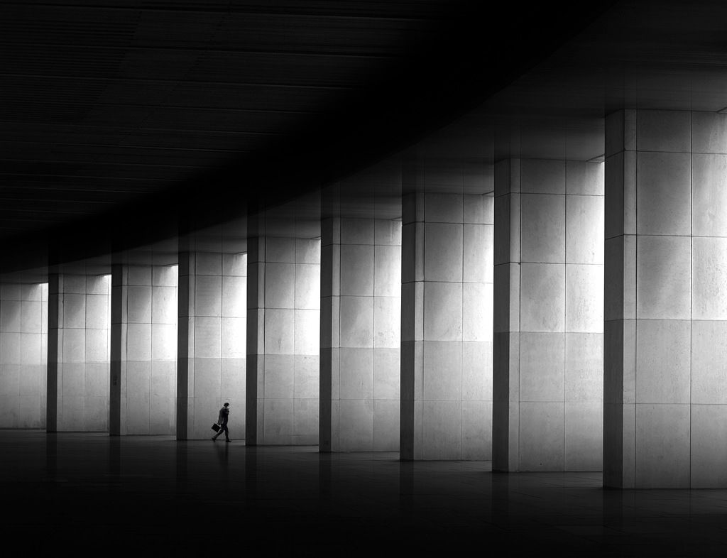 A lone figure with a briefcase walking through a sunlit colonnade of towering stone pillars, rendered in dramatic black and white