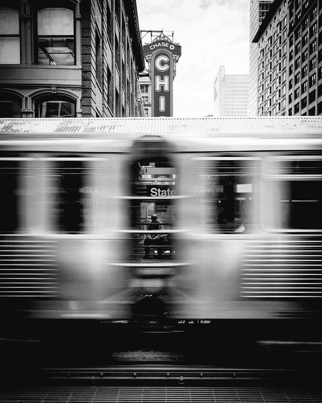Black-and-white street photograph of a CTA L train streaking past the State Street station in motion blur, with the Chicago Theatre marquee rising over downtown buildings