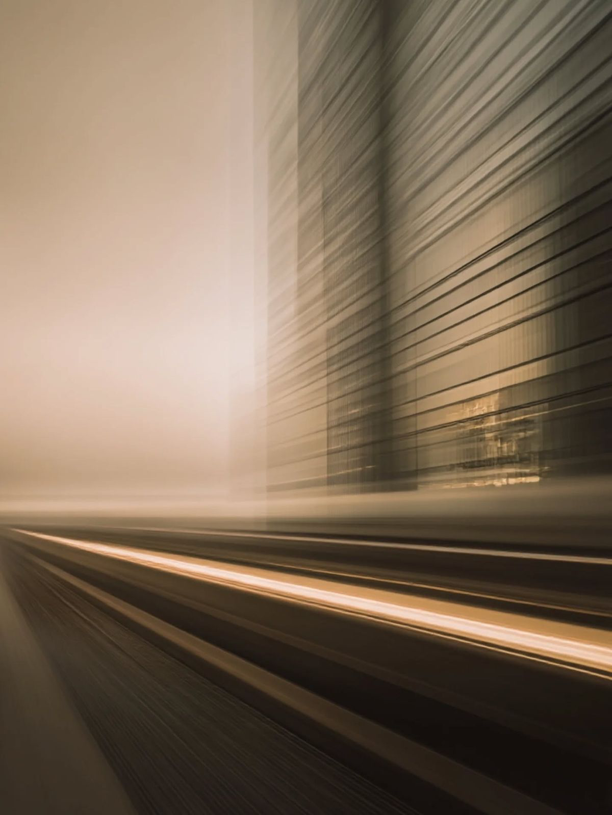A warm, sepia-toned motion-blur photograph of a modern glass high-rise streaking past with light trails along tracks in the foreground